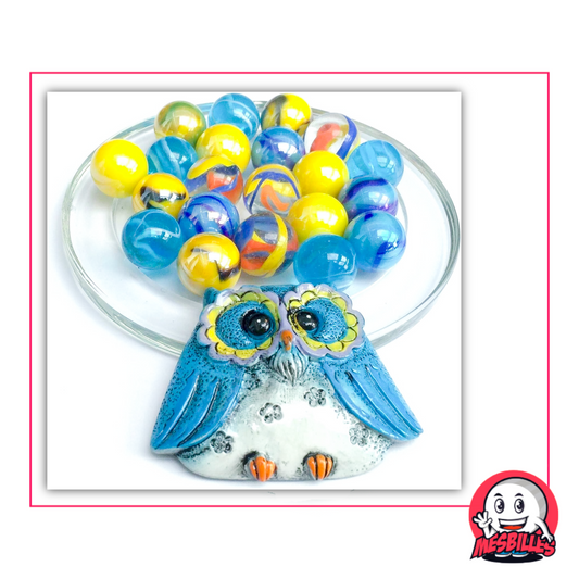 Búho azul de cerámica acompañado de 20 canicas de 16 mm de cristal azul, amarillo, transparente y marmolado, colocadas sobre una copa de cristal con marco rosa y el logotipo de MisCanicas.